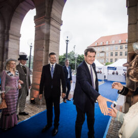 Das Bild zeigt eine Gruppe von Menschen auf einem blauen Teppich unter einem Torbogen, darunter Personen in historischer Kleidung und zwei Männer in modernen Anzügen. Einer der Männer in Anzug schüttelt einer Person in historischer Kleidung die Hand. Im Hintergrund sind ein Gebäude mit vielen Fenstern sowie Zelte oder Stände zu sehen, was auf eine Veranstaltung mit historischem Bezug hinweist.