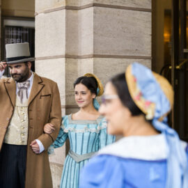 Das Bild zeigt eine Gruppe von Menschen in historischen Kostümen vor einem Gebäude mit steinernen Säulen. Im Mittelpunkt steht ein Mann mit braunem Mantel, beigem Westenanzug und Zylinder, der den Arm einer Frau in einem blauen Kleid mit Spitzenbesatz hält. Weitere Personen in historischer Kleidung sind im Hintergrund zu sehen.