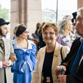 Das Bild zeigt eine Gruppe von Menschen in historischer oder nostalgischer Kleidung. Im Vordergrund sind fünf Personen zu sehen, darunter eine Person mit Melone, hellem Anzug und weißen Handschuhen sowie eine weitere in einem blauen Kleid mit weißen Akzenten und Kopftuch. Weitere Personen tragen elegante Kleidung mit Accessoires wie Perlen, Broschen oder einem Gehstock. Im Hintergrund befinden sich weitere Menschen in ähnlicher historischer Kleidung, was auf eine Veranstaltung mit historischem Modethema hinweist.