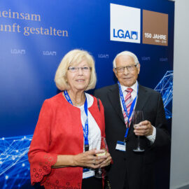 Das Bild zeigt zwei Personen in formeller Kleidung, die vor einem blauen Hintergrund mit der Aufschrift „Gemeinsam Zukunft gestalten“. Beide tragen Namensschilder an einem Band um den Hals und Weingläser.