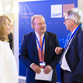 Das Bild zeigt drei Personen im Gespräch bei einer professionellen oder formellen Veranstaltung. Sie stehen vor einem blauen Hintergrund mit der Aufschrift „Gemeinsam Zukunft gestalten“ und dem Logo „LGA“. Alle tragen Businesskleidung und Namensschilder.