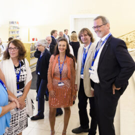 Das Bild zeigt eine Gruppe von Menschen, die sich in einem formellen oder professionellen Rahmen unterhalten. Alle tragen Namensschilder an Bändern, was auf eine Veranstaltung hinweist. Im Hintergrund sind eine Treppe mit verzierten Geländern, Regale mit Büchern oder Materialien sowie Türen zu weiteren Räumen zu sehen.