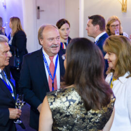 Mehrere Personen stehen in einem Innenraum bei einer formellen Veranstaltung oder Konferenz. Sie tragen Businesskleidung und Namensschilder an blauen Bändern, unterhalten sich und halten teilweise Getränke in der Hand.