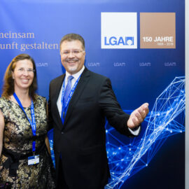 Zwei Personen stehen vor einem blauen Hintergrund mit Logos und einem Jubiläumshinweis. Beide tragen formelle Kleidung und Namensschilder an Bändern um den Hals.