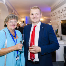Zwei Personen stehen in einem hell erleuchteten Raum und halten Getränke in der Hand. Die linke Person trägt ein hellblaues Outfit, die rechte einen dunkelblauen Anzug mit weißem Hemd und roter Krawatte. Im Hintergrund sind weitere Menschen, Möbel sowie ein Bildschirm und ein Willkommensschild an der Wand zu sehen.
