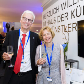 Zwei Personen stehen nebeneinander bei einer Veranstaltung und halten jeweils ein Glas Wein in der Hand. Beide tragen Namensschilder an blauen Bändern. Im Hintergrund ist eine Wand mit einem Willkommensschriftzug.