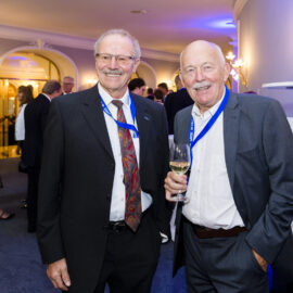 Bei einer gesellschaftlichen Veranstaltung stehen zwei Personen im Vordergrund, beide in Anzügen und mit blauen Namensbändern. Eine der Personen hält ein Glas Weißwein. Im Hintergrund unterhalten sich weitere Gäste in einem elegant gestalteten Innenraum mit weicher Beleuchtung und dekorativen Elementen wie Wandlampen und Rundbögen.