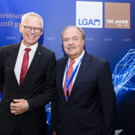 Zwei Personen in Businesskleidung stehen vor einem blauen Hintergrund mit grafischen Elementen aus Linien und Knotenpunkten. Der Hintergrund zeigt Logos und einen Jubiläumshinweis.