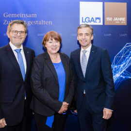 Drei Personen stehen vor einem blauen Hintergrund mit Logos und einem Jubiläumshinweis. Der Hintergrund zeigt den Schriftzug einer gemeinsamen Zukunftsgestaltung.