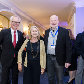 Drei Personen stehen im Vordergrund einer Veranstaltung in einem eleganten Raum mit stilvollen Leuchten und Rundbögen. Weitere Gäste sind im Hintergrund zu sehen, einige tragen Namensschilder an Bändern. Die Kleidung der Anwesenden ist formell, was auf ein Event hinweist.
