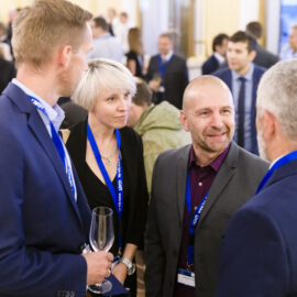 Mehrere Personen befinden sich bei einer beruflichen oder Netzwerkveranstaltung und unterhalten sich miteinander. Sie tragen Namensschilder an blauen Bändern, eine Person hält ein leeres Weinglas. Im Hintergrund sind weitere Gäste in Gespräche vertieft, was auf eine gesellige Atmosphäre hinweist.