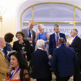 Mehrere Personen in formeller Kleidung stehen in einem eleganten Innenraum mit warmem Licht und dekorativen Elementen wie Kronleuchtern. Im Hintergrund ist ein großer Rundbogen mit goldenen Verzierungen und ein teilweise sichtbares Willkommensschild zu sehen. Einige Gäste halten Getränke und unterhalten sich.