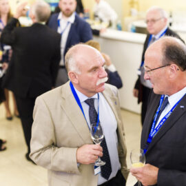 Mehrere Personen befinden sich bei einer gesellschaftlichen oder beruflichen Veranstaltung und tragen Businesskleidung sowie Namensschilder an blauen Bändern. Einige Gäste halten Getränke in der Hand und unterhalten sich.