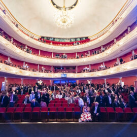 Das Bild zeigt das Innere eines prunkvollen Theaters oder Opernhauses mit mehreren Rängen und einem großen Kronleuchter an der Decke. Die Zuschauer sitzen in roten Stühlen, einige stehen oder gehen umher, was auf eine Pause oder den Beginn der Veranstaltung hindeutet. Die Architektur ist reich verziert mit weißen und goldenen Elementen.