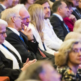 Mehrere Personen sitzen in einem Auditorium oder Konferenzraum und blicken nach vorn, als würden sie einer Präsentation oder einem Vortrag folgen. Einige tragen Namensschilder oder Bänder um den Hals.