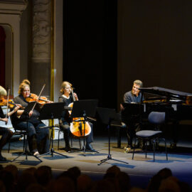Vier Musikerinnen und Musiker treten auf einer Bühne in einem klassisch dekorierten Konzertsaal auf. Zu sehen sind zwei Violinistinnen oder Violinisten, eine Cellistin oder ein Cellist sowie eine Person am Klavier, alle mit Notenständern vor sich. Im unteren Bildbereich ist das Publikum zu erkennen, was auf ein Live-Konzert hinweist.