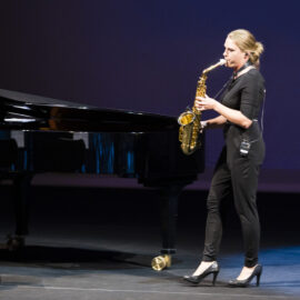 Eine Person steht auf einer Bühne und spielt Saxophon, gekleidet in Schwarz und mit hochhackigen Schuhen. Im Hintergrund befinden sich ein Flügel und ein leerer Stuhl. Die Bühne ist dunkel gehalten, das Licht richtet sich auf die Musikerin.
