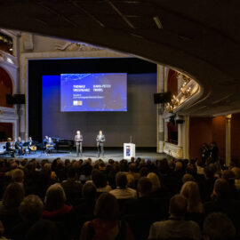 In einem prunkvoll dekorierten Theater mit Kronleuchtern und Balkonen findet eine formelle Veranstaltung statt. Auf der Bühne stehen zwei Personen vor einer großen Leinwand, während links eine kleine Musikgruppe spielt. Das Publikum sitzt und blickt zur Bühne.