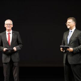 Zwei Personen stehen auf einer Bühne und tragen formelle Kleidung. Der Hintergrund ist dunkel.