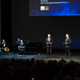 Auf einer Bühne stehen zwei Männer in Anzügen, einer mit Mikrofon, der andere mit einem Tablet. Links daneben sitzt ein Ensemble mit verschiedenen Musikinstrumenten wie Klavier, Violine, Cello und Klarinette, im Hintergrund ist eine große Leinwand mit Logos und Text zu sehen, davor das Publikum.