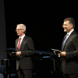 Zwei Personen stehen auf einer Bühne und tragen formelle Kleidung, beide halten Papiere in der Hand. Im Hintergrund ist eine weitere Person teilweise sichtbar, möglicherweise mit einem Musikinstrument. Die dunkle Umgebung deutet auf eine Veranstaltung oder Aufführung in einem Innenraum hin.