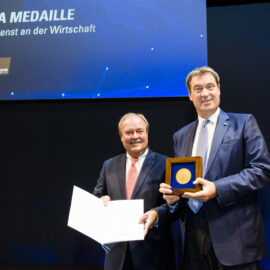 Zwei Personen stehen auf einer Bühne, eine hält eine geöffnete Urkunde oder ein Dokument, die andere eine gerahmte Medaille. Im Hintergrund ist eine große Leinwand mit einem Schriftzug und Logos zu sehen.
