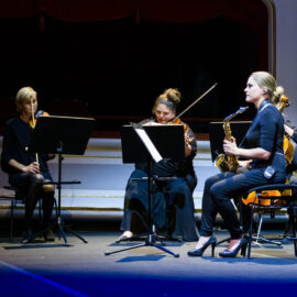 Drei Musiker sitzen auf einer Bühne und spielen verschiedene Instrumente: links ein Streichinstrument, in der Mitte ein Cello und rechts ein Saxophon. Vor ihnen stehen Notenständer, die Bühne ist in dunklem Licht mit blauen Akzenten beleuchtet.