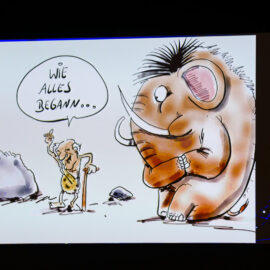 Das Bild zeigt eine Cartoon-Zeichnung mit einem älteren Höhlenmenschen, der sich mit einem Stock an einen großen wolligen Mammut wendet. Im Hintergrund sind Felsen zu sehen, die Szene spielt in einer prähistorischen Umgebung.