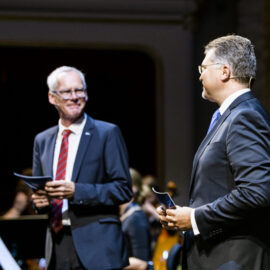 Zwei Personen in Anzügen stehen auf einer Bühne, beide halten Papiere in der Hand. Im Hintergrund sind Musikinstrumente und weitere Personen zu sehen, was auf eine Festveranstaltung hinweist.