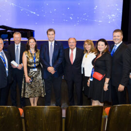Zehn Personen auf einer Bühne, alle tragen formelle Kleidung wie Anzüge, Krawatten oder Businesskleidung. Im Hintergrund ist eine blaue Leinwand mit weißen Lichtmustern zu sehen, links steht ein Flügel. Alle Personen tragen Namensschilder an einem Band um den Hals, was auf eine Veranstaltung hinweist.