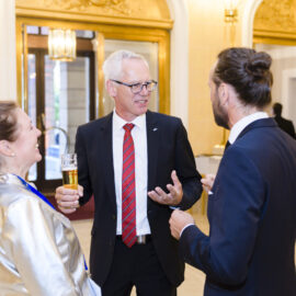 Drei Personen unterhalten sich bei einer formellen Veranstaltung, die mittlere Person hält ein Glas Bier und trägt einen schwarzen Anzug mit rot gestreifter Krawatte. Links steht eine Person in einer glänzenden silbernen Jacke, rechts eine Person im dunklen Anzug. Der Hintergrund zeigt eine prunkvolle Umgebung mit goldenen Verzierungen und Kronleuchtern, weitere Gäste sind im Raum verteilt.