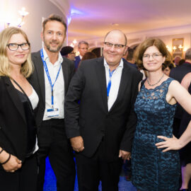 Eine Gruppe von Personen in formeller Kleidung befindet sich bei einer gesellschaftlichen oder beruflichen Veranstaltung in einem Innenraum. Im Hintergrund sind weitere Gäste und stimmungsvolle Beleuchtung zu sehen, was auf eine festliche oder formelle Atmosphäre hinweist.