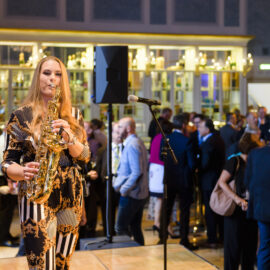 Das Bild zeigt eine gesellschaftliche Veranstaltung in einem großen, elegant dekorierten Raum mit warmem Licht. Im Vordergrund spielt eine Person Saxophon, im Hintergrund unterhalten sich mehrere Gäste an Tischen mit Getränken und Speisen. Ein Mikrofonständer und ein Lautsprecher sind ebenfalls sichtbar.