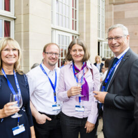 Fünf Personen stehen bei einer formellen oder beruflichen Veranstaltung zusammen, alle tragen Namensschilder an blauen Bändern um den Hals. Drei von ihnen halten Weingläser in der Hand. Im Hintergrund sind große Fenster und ein Gebäude aus Stein zu sehen.