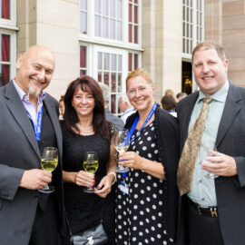 Vier Personen stehen bei einer gesellschaftlichen oder formellen Veranstaltung zusammen, alle tragen elegante Kleidung und halten ein Glas Weißwein in der Hand. Im Hintergrund sind große Fenster und Steinwände zu sehen, was auf einen stilvollen Veranstaltungsort hinweist. Weitere Gäste unterhalten sich im Hintergrund.