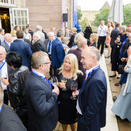 Eine große Gruppe von Personen hat sich bei einer Veranstaltung im Freien versammelt, viele tragen formelle Kleidung und halten Weingläser in der Hand. Die meisten haben Namensschilder an blauen Bändern um den Hals. Der Veranstaltungsort scheint eine Terrasse oder ein Hof neben einem Gebäude mit großen Fenstern und Steinwänden zu sein.