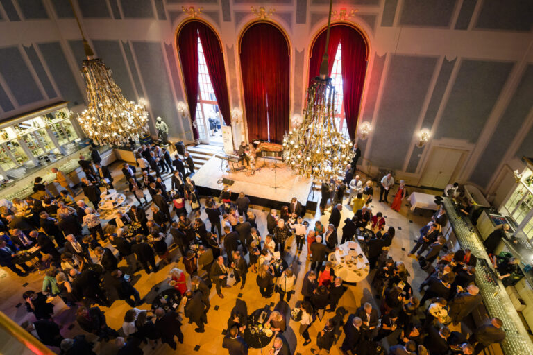Das Bild zeigt einen großen, eleganten Saal mit hohen Decken, prunkvollen Kronleuchtern und großen roten Vorhängen vor hohen Fenstern. In der Mitte befindet sich eine Bühne, auf der Musiker auftreten. Die Gäste tragen formelle Kleidung und stehen in Gruppen an Tischen, unterhalten sich und genießen die Veranstaltung.