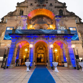 Der Eingang eines prunkvollen Gebäudes ist in blauem und gelbem Licht beleuchtet. Die Architektur zeigt große Rundbögen, verzierte Details und Statuen. Ein blauer Teppich führt zum Eingang, gesäumt von kleinen runden Tischen mit weißen Tischdecken.