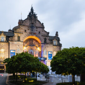 Ein historisches Gebäude mit aufwendiger Architektur und großem Rundbogen-Eingang ist beleuchtet und hebt so seine Details hervor. Davor befinden sich mehrere Bäume sowie ein Außenbereich mit Tischen und Stühlen, was auf eine Veranstaltung hinweist. Am Gebäude sind zudem Banner angebracht, der Himmel ist bewölkt.