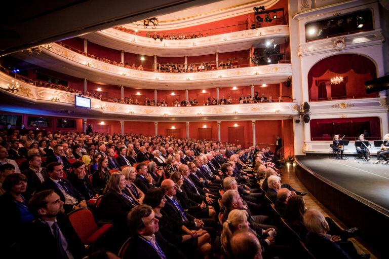 Das Bild zeigt ein prunkvoll gestaltetes Theater mit mehreren Balkonen, in dem eine große Anzahl von Menschen Platz genommen hat. Der Saal ist gut beleuchtet, und das Publikum scheint einer Aufführung oder Veranstaltung auf der Bühne aufmerksam zu folgen. Die Architektur des Theaters ist aufwendig und detailreich gestaltet.