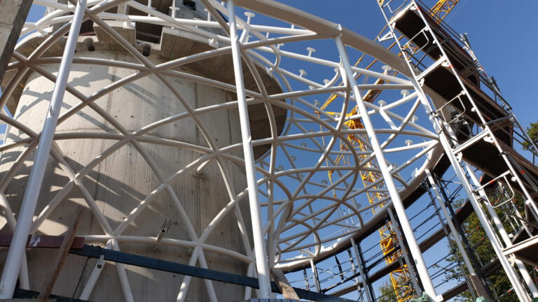 Baustelle mit großer zylindrischer Betonstruktur, umgeben von einem komplexen weißen Metallgerüst; im Hintergrund sind Gerüste und ein gelber Kran vor blauem Himmel zu sehen.