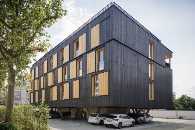 Modernes, mehrstöckiges Gebäude mit dunkler Fassade und hellen hölzernen Fensterläden, auf Stelzen errichtet mit einem offenen Bereich darunter, in dem mehrere Autos parken; links im Bild befinden sich ein Baum und etwas Grünfläche.