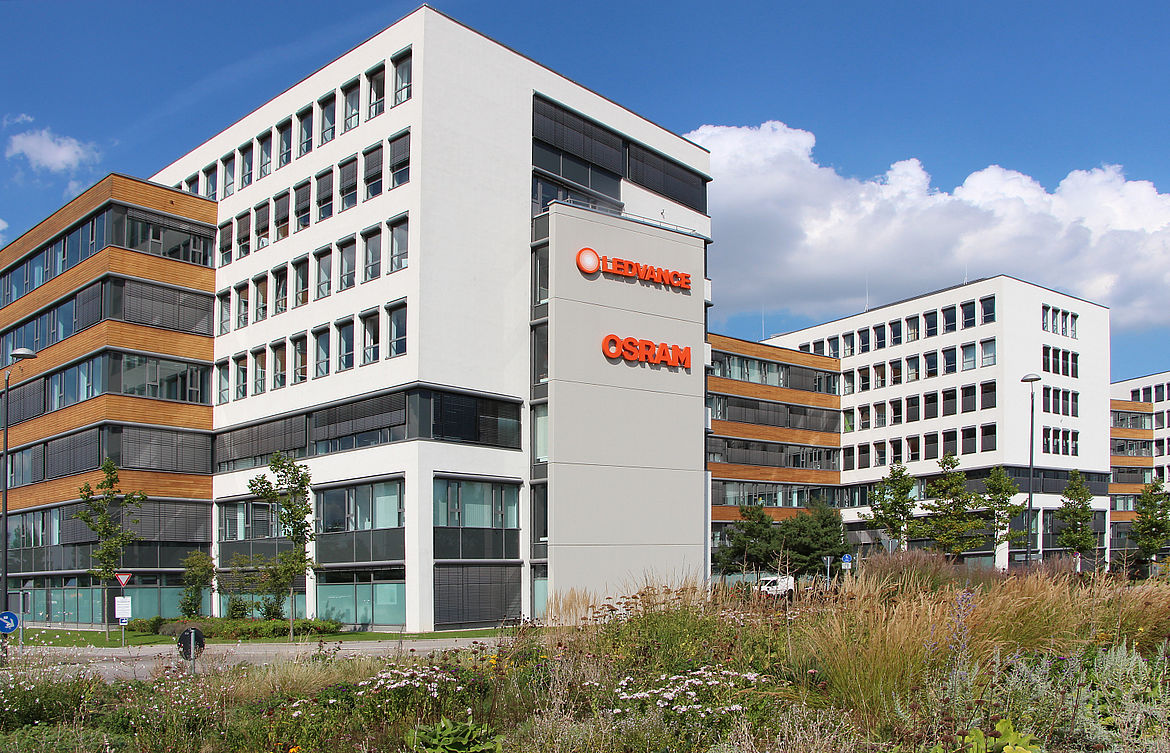 Modernes Bürogebäude-Ensemble mit mehreren Gebäuden, im Vordergrund ein Hauptgebäude mit der Aufschrift „LEDVANCE OSRAM“ in roten Buchstaben, umgeben von Grünflächen und gepflegter Landschaftsgestaltung.