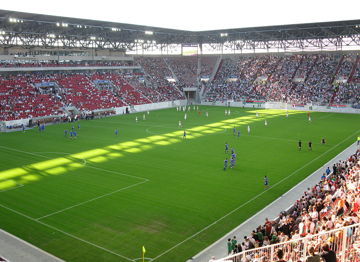 Fußballstadion mit roten Sitzplätzen, das größtenteils mit Zuschauern gefüllt ist; auf dem Spielfeld findet ein laufendes Spiel zwischen Mannschaften in blauen und weißen Trikots statt, mit Schattenwurf durch die Stadionüberdachung.