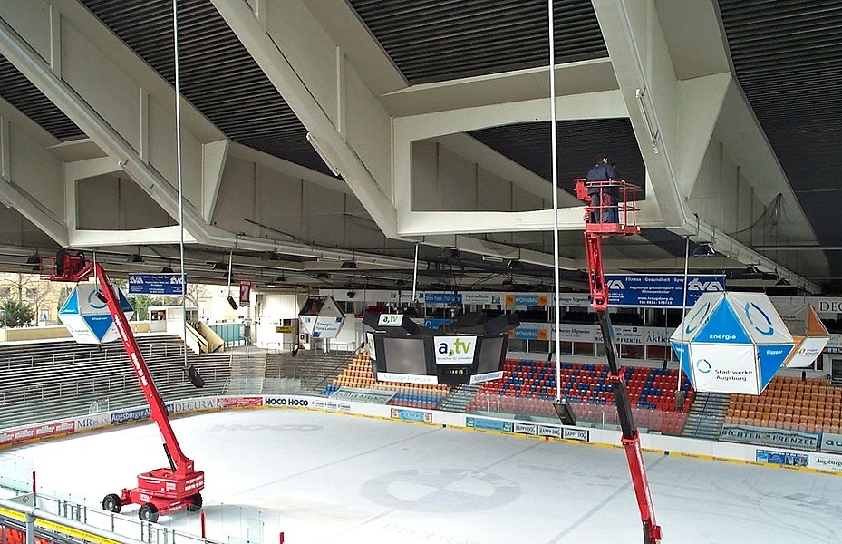 Zwei rote Hebebühnen stehen in einer Eishalle und werden für Wartungsarbeiten an der Decke eingesetzt; im Hintergrund sind bunte Tribünen, ein zentraler Videowürfel und Werbebanner rund um das Spielfeld zu sehen.