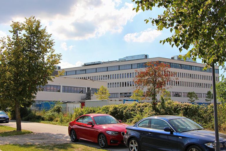 Ein großes, modernes Gebäude mit mehreren Etagen und zahlreichen Fenstern steht im Hintergrund; im Vordergrund sind ein rotes und ein blaues Auto geparkt, umgeben von Bäumen, Grünflächen und einem teils bewölkten Himmel.