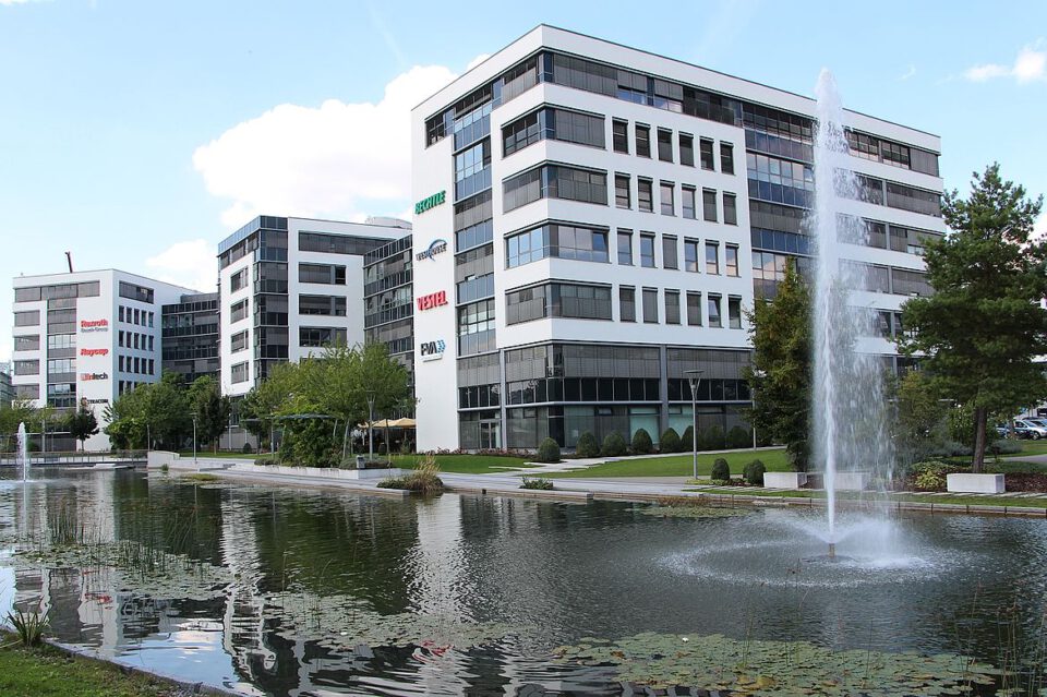 Ein modernes Bürokomplex mit mehreren mehrstöckigen Gebäuden umgibt einen großen Teich mit Springbrunnen, umgeben von Bäumen und Grünflächen; auf den Gebäuden sind Firmenlogos.