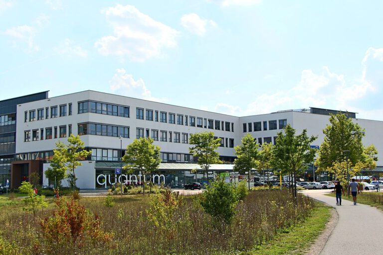 Modernes, mehrstöckiges Gebäude mit großen Fenstern und dem Schriftzug „quantum“ an der Fassade, umgeben von einer begrünten Anlage mit Gehweg, Bäumen und parkenden Autos unter teils bewölktem Himmel.