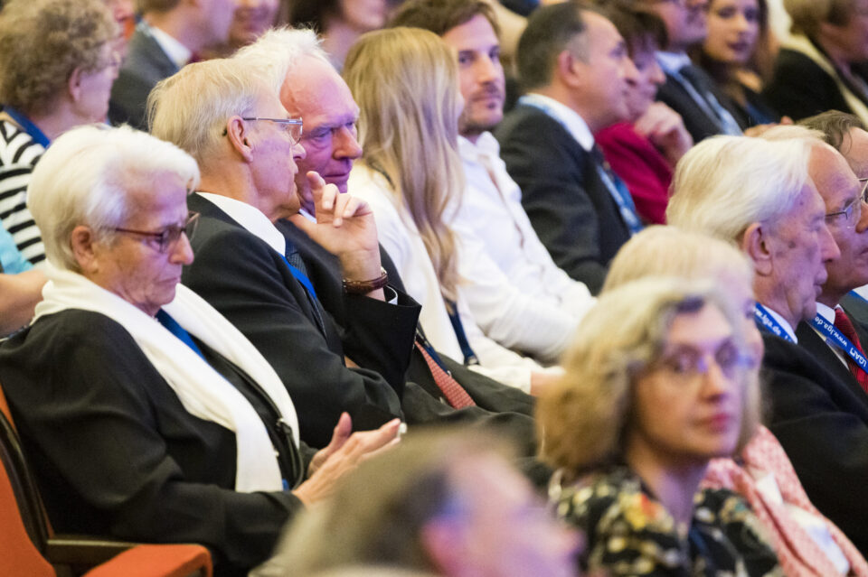 Mehrere Personen sitzen in einem Auditorium oder Konferenzraum und blicken nach vorn, als würden sie einer Präsentation oder einem Vortrag folgen. Einige tragen Namensschilder oder Bänder um den Hals.
