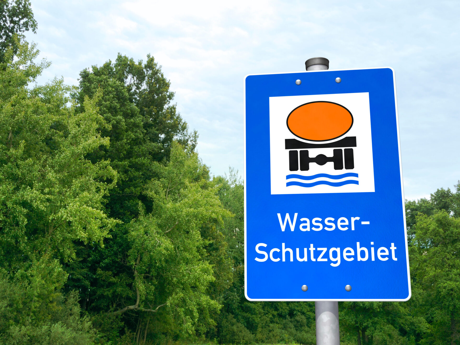 Das Bild zeigt ein blaues Hinweisschild mit einem weißen Quadrat, in dem ein orangefarbener Kreis, schwarze Formen und blaue Wellenlinien abgebildet sind, die Wasser und Infrastruktur symbolisieren. Unter der Grafik steht der Schriftzug Wasser-Schutzgebiet. Im Hintergrund sind grüne Bäume und Pflanzen zu sehen.